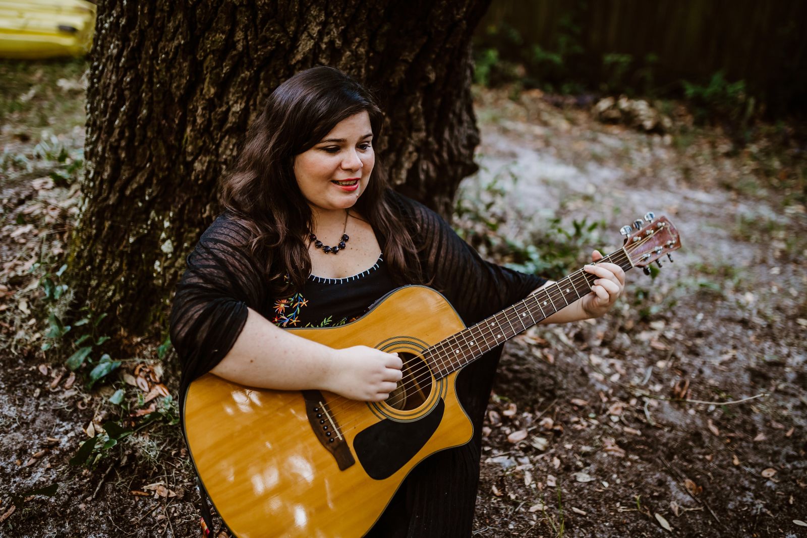 Photo of musician Ashley Feller sitting on ground under a large tree playing guitar