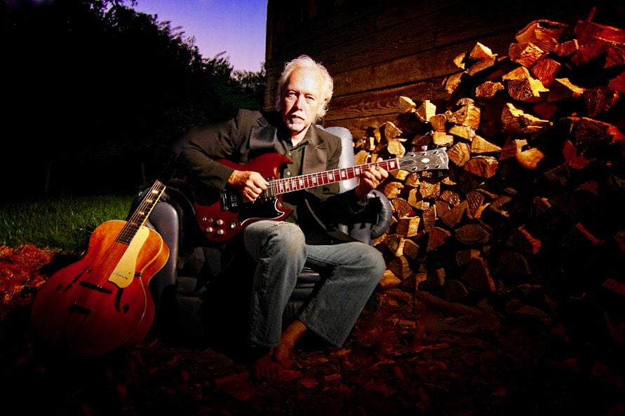 A photograph of a man sitting near a pile of wood at sunset, playing a guitar with another guitar nearby.
